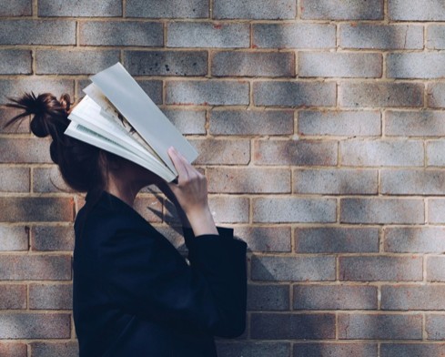 Woman covering her face with an open book against a brick wall